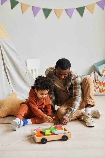 Vertical portrait of black father and son playing with toys together in cute kids room interior