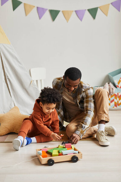 Вертикальный портрет черного отца и сына, играющих вместе с игрушками в интерьере детской комнаты