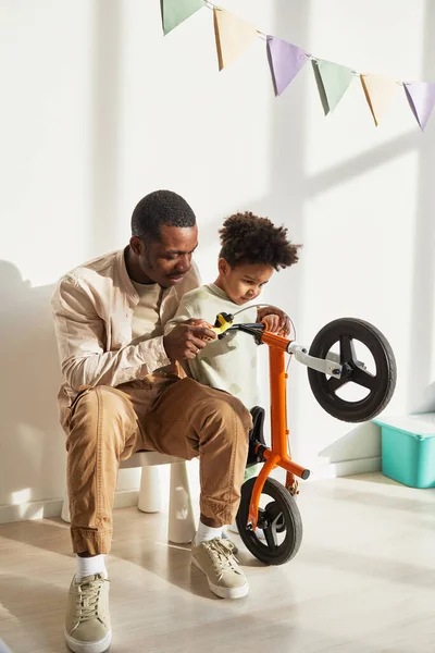 Vertical portrait of loving black father teaching little boy riding balance bike at home in sunlight