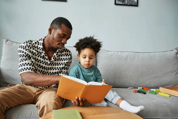 Portrait of black father and son reading books together while sitting on couch at home, copy space