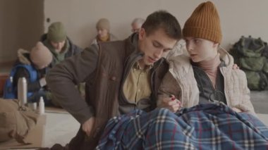 Küçük oğlu ve yetişkin çifti olan genç bir kadın da dahil olmak üzere diğer mültecilerle birlikte soğuk sığınma evinde kalırken güç bankasını cep telefonlarını şarj etmek için kullanan mültecilerin orta derecede yavaşlaması.