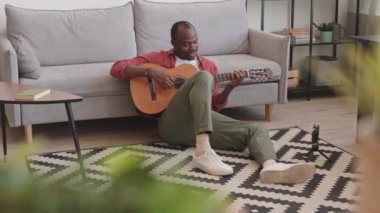 Kırmızı tişörtlü genç siyahi adam, oturma odasında kız arkadaşıyla birlikte gitar çalarken videosunu kaydediyor. Müzik dinliyor ve akıllı telefon kamerasına el sallıyor.