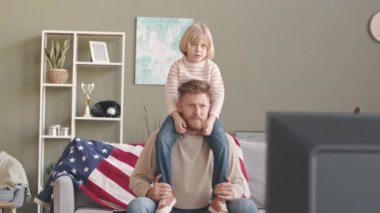 Oturma odasındaki kanepede birlikte muhteşem bir spor maçı izlerken küçük oğlunu omuzlarında taşıyan sakallı, beyaz bir adam.