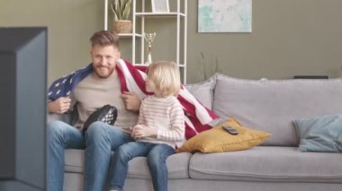 Beyaz bir adam ve küçük oğlu, oturma odasında Amerikan bayrağı ve beyzbol ekipmanıyla kanepede oturup, takımlarının kazanmasını kutlarken televizyonda beyzbol maçı izliyorlar.