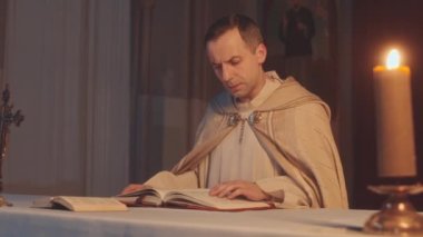 Beyaz alb ve pelerinli Kafkasyalı rahibin orta yavaşlığında kutsal İncil okurken mihrapta koyu Katolik kilisesinde mum yanıyordu.