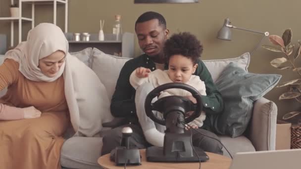 美国黑人穆斯林的父亲和3岁的儿子在家里用游戏方向盘玩电脑游戏 戴头巾的男孩妈妈坐在旁边看着 — 图库视频影像