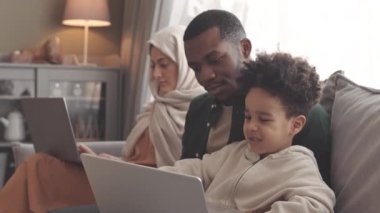Yavaş yavaş Afrikalı Amerikalı Müslüman bir baba ve yeni yürümeye başlayan oğlu dizüstü bilgisayardan çizgi film seyrederken karısı başörtüsü takmış kanepenin yanında dizüstü bilgisayarda çalışıyor.