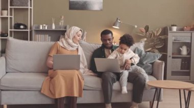 Üç kişilik modern, ırklar arası Müslüman bir aile oturma odasındaki kanepede iki dizüstü bilgisayarla oturuyor. Eşi ve küçük oğlu çizgi film izlerken türbanlı bir kadın çalışıyor.