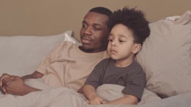 Yavru oğluyla akşam televizyon izlerken kanalı değiştiren, yumuşak battaniyenin altında birlikte uzanan Afrikalı Amerikalı babanın yavaşlığı.