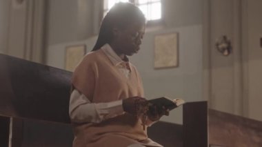 Ellerinde tespih boncuklarıyla Katolik kilisesindeki ahşap bankta oturan ve dua eden genç Afrikalı Amerikalı kadın cemaat mensubu orta yavaş.