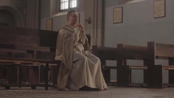 身穿白衣的白人牧师慢吞吞地坐在天主教堂的木制长椅上 闭上眼睛 双手紧握着 向上帝祈祷 — 图库视频影像