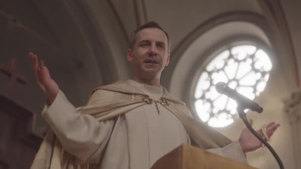 ミサ中に教区に説教をしながら 白の投資の牧師の低角度の低速のジェスチャー カトリック教会のマイクでTribuuneに立って — ストック動画