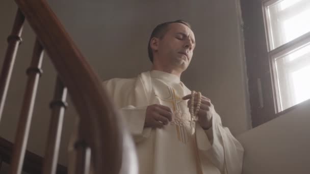 カトリック教会の木製の階段の上に窓のそばに立っている間 バラのビーズを保持し 神に祈る白い子羊の成熟した司祭の減速 — ストック動画