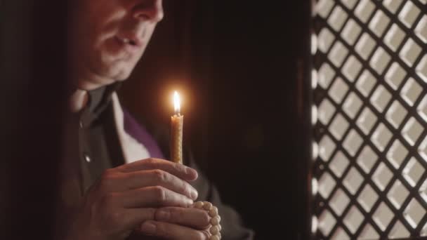 カトリック教会の暗い告白ブースに座っている間にろうそくを吹いて黒と紫のローブを身に着けている成熟した司祭のスロー — ストック動画