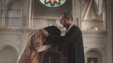 Siyah cüppeli olgun Kafkasyalı rahibin orta boy ağırlığı Katolik kilisesinde genç Afrikalı Amerikalı kadını kutsuyor.