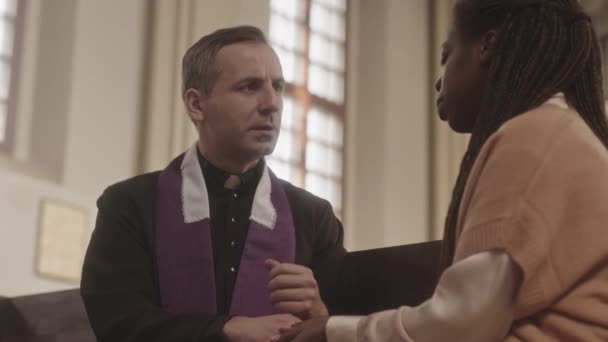 ウエストアップ遅いです成熟した白人聖職者で黒と紫のローブと若いアフリカ系アメリカ人女性の教区が会話を持ちます 手を握ってカトリック教会で木製のPewに座って — ストック動画