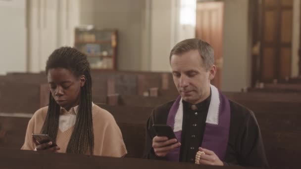 多民族の司祭と若い女性の教区のスローモは 彼らのスマートフォンを使用してカトリック教会の木製のベンチに隣に座っています — ストック動画