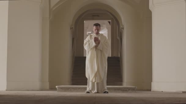 教会の入り口にある2本の背の高いベージュの柱の間に立つ長い白い衣を着た司祭の完全な長さのスローモと神に祈る — ストック動画