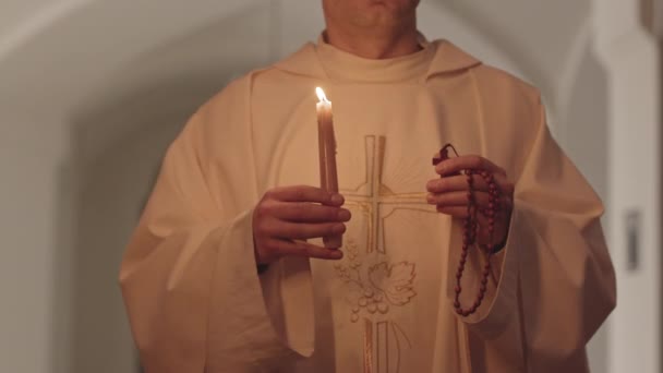白い衣に金の十字架の刺繍が片手に燃えるろうそくとバラ色のビーズを持つ教会に沿って歩くと 認識できない司祭のミッドセクションのスローモ別の — ストック動画