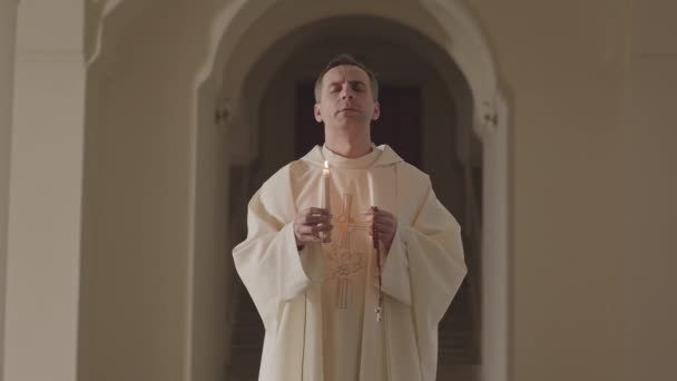 カトリック教会の廊下に立つ 燃えるろうそくとバラ色のビーズを保持し 神に祈る長い白いアルバムで成熟した白人の司祭の中低速 — ストック動画
