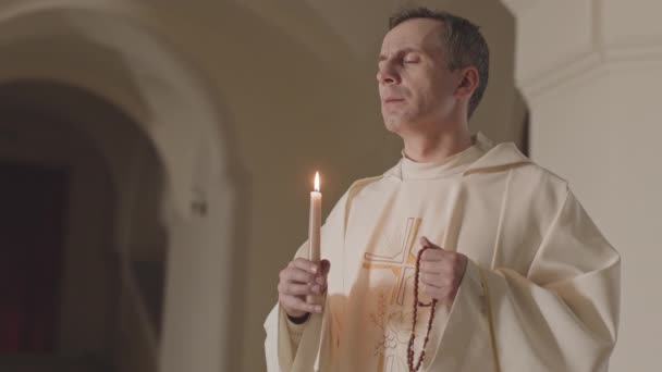お祈りの後にろうそくを吹いて 手にバラ色のビーズを保持長い白いローブで成熟した白人の司祭の中低速 — ストック動画