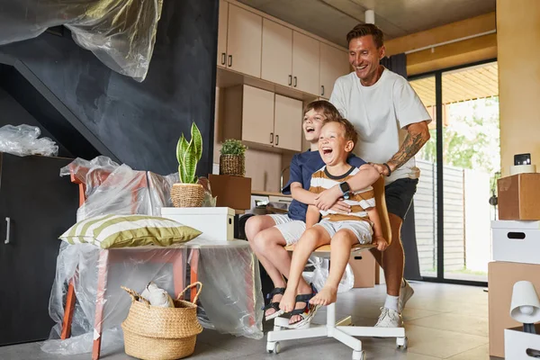 幸せな家族の完全な長さの肖像画新しい家に移り住み 屈託のない父は子供たちと一緒にホールで椅子に乗って遊ぶ — ストック写真