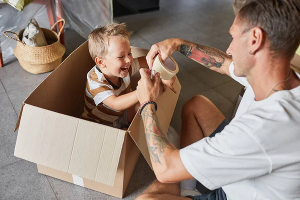 家族の引っ越しの家のために梱包しながら 段ボール箱の中でかわいい男の子と遊ぶ幸せな父親の肖像画 — ストック写真