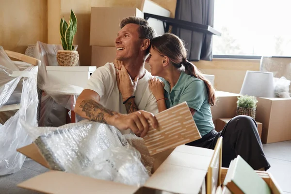 Portrait of happy tattooed couple unpacking boxes sitting on floor while moving into new house, copy space