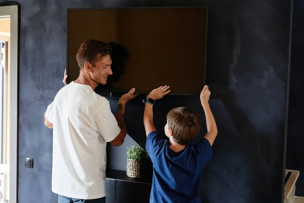 Minimal back view portrait of smiling father and son hanging TV on wall together while moving to new home