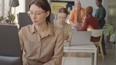 Modern dijital pazarlama ajansında arka planda iletişim kuran çeşitli çalışma arkadaşlarıyla birlikte çalışırken, kablosuz klavyede yazan Kafkasyalı genç bir kadının orta yavaşlığı.