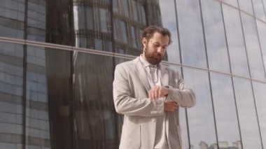 Sakallı, beyaz, akıllı iş adamı yavaş yavaş uzun cam ofis binasını terk ediyor ve güneşli bir günde şehir merkezinde dışarıda beklerken akıllı telefonuyla telefon görüşmesi yapıyor.