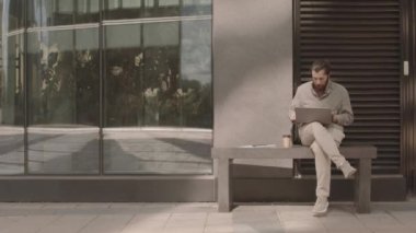 Sakallı genç Kafkasyalı iş adamı elinde kahve ile modern ofis binasının yanındaki bankta oturuyor bilgisayarla çalışıyor, belgeleri inceliyor ve telefon görüşmeleri yapıyor.