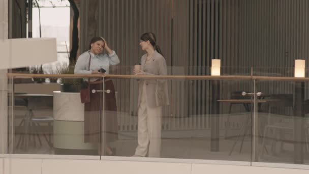 現代のビジネスセンターでガラスの手すりに立っている間 エレガントなスマートカジュアルウェアの話で2人の若い多民族女性の完全な長さの低速 — ストック動画