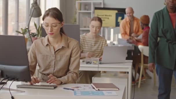 在现代数字营销代理中 一群年轻的 多样化的人在一起工作 年轻的白种人妇女戴着眼镜 坐在电脑前检查文件 前景光明 — 图库视频影像