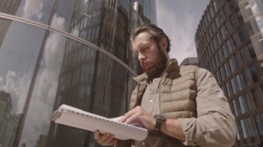 Şehir merkezinde güneşli bir günde modern cam ofis binalarının altında dikilirken yorganlı yelek giymiş, iş belgelerine bakarken orta açılı bir fotoğraf.