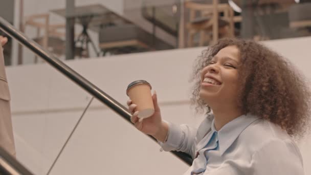 若いですCurvyアフリカ系アメリカ人女性の遅いですコーヒーカップとともに彼女の女性の友人と話をしながら上にエスカレーター ショッピングで一緒に大きな現代的なデパート — ストック動画