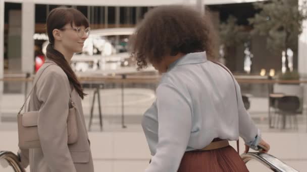 两名多种族的年轻成功女性慢吞吞地走下现代购物中心的自动扶梯 与人交谈 — 图库视频影像