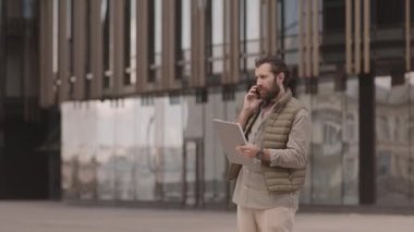 Şehir merkezindeki iş belgeleri ve laptopla birlikte akıllı telefon görüşmeleri ve video sohbetlerinin orta boy sakallı girişimci fotoğrafı.