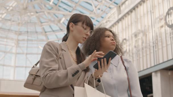 スマートフォンを見回し閲覧する2人の若い幸せな多様な女性の低角度中低速 大規模な近代的なデパートで買い物袋と立って — ストック動画