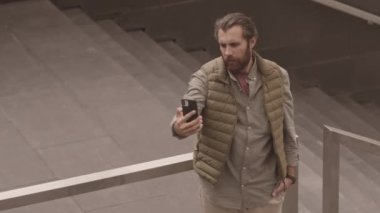 Kafkas sakallı bir adamın arkadaşlarıyla ya da meslektaşlarıyla görüntülü konuşma yaparken akıllı telefon kamerasına el salladığı, dışarıda korkuluklara yaslandığı yukarıdan çekilmiş bir fotoğraf.