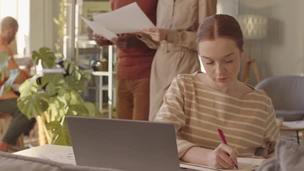 若い白人女性の中低速コピーブックでノートを取りながら 成功したデジタルマーケティング代理店の明るい近代的なオフィスのデスクに座ってノートPc上で市場分析を行う — ストック動画