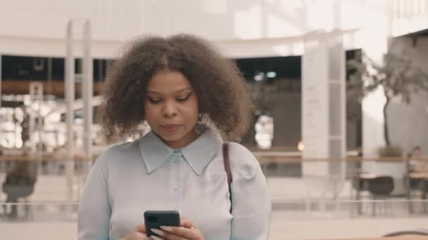 中低速肖像画の若いカービーアフリカ系アメリカ人女性のライトブルーのシャツスクロールスマートフォンその後笑顔でカメラスタンドで新しいショッピングセンターの1階 — ストック動画