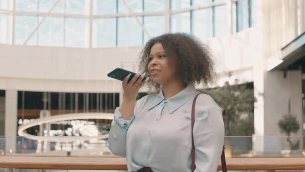 中低速の若いアフリカ系アメリカ人女性のライトブルーのシャツのスピーカーフォン録音の音声メッセージで話す現代のショッピングセンター — ストック動画
