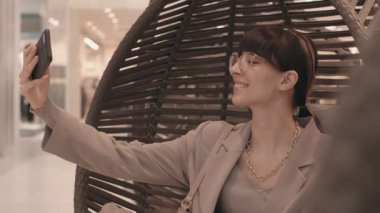 Modern alışveriş merkezindeki asılı yuva sandalyesinde oturmuş, akıllı telefonunda fotoğraf çeken, şık giysili ve gözlüklü esmer bir kadının orta boy yavaşlığı.