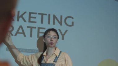 Genç kadın SMM uzmanının, güncel pazarlama eğilimleri ve bir grup insana başarılı stratejiler hakkında sunum yapması, karanlık odada ekran projektörüne karşı durması,