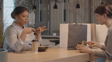 Afro-Amerikalı genç bir kadının akıllı telefonuyla öğle yemeğinin fotoğraflarını çekmesi ve alışveriş yaptıktan sonra birlikte rahat bir kafede dinlenirken bayan arkadaşına göstermesi.