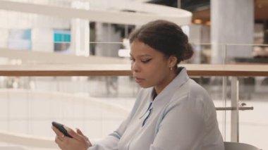 Orta yaşlı, kıvrımlı, Afro-Amerikan iş kadını açık mavi gömlekli, akıllı telefon kullanan ve modern mağazadaki kafede oturmuş gülümseyen.