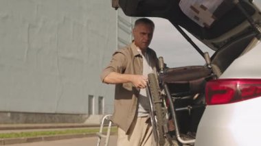 Engelli yetişkin bir beyaz adam dışarıda park etmiş bir arabanın bagajından tekerlekli sandalye alıyor.