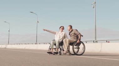 Tekerlekli sandalyedeki olumlu beyaz çift yaz gününü dışarıda sohbet ederek geçiriyor. Bir kadın kocasını işaret ediyor.