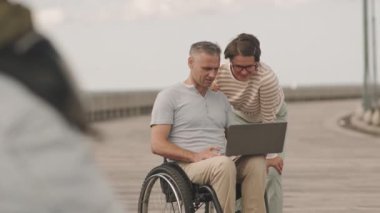 Tekerlekli sandalyedeki olgun beyaz adam ve karısı dışarıda rıhtımda vakit geçirirken bilgisayarlarında arkadaşlarıyla görüntülü sohbet ediyorlar.